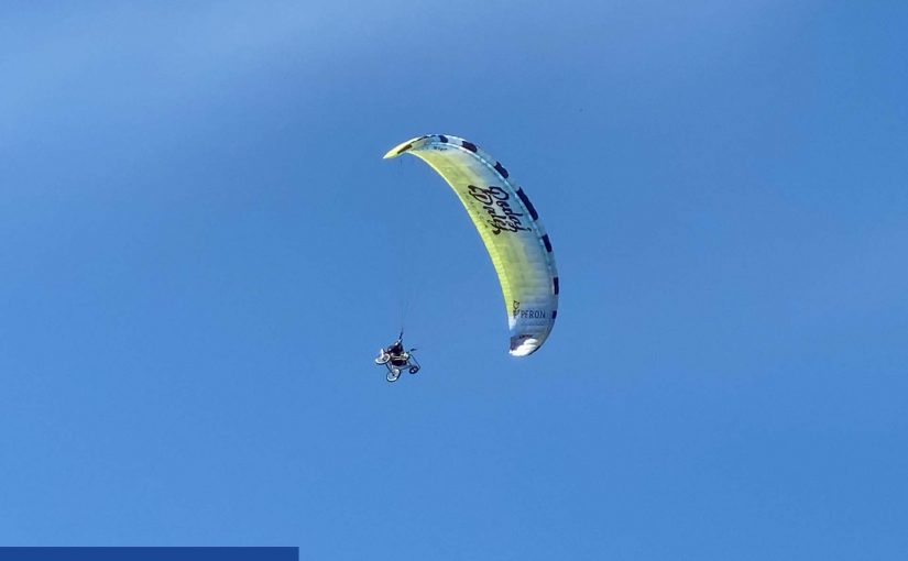 Na tle nieba widać biało- żółte skrzydło paralotni z wyraźnym logo Fundacji PODAJ DALEJ. Pilot paralotni siedzi na wózku.