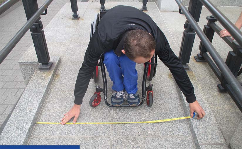 Zdjęcie wykonane na podjeździe dla osób z niepełnosprawnością. W centrum przodem do nas stoi mężczyzna na wózku, który schyla się (nie widać jego twarzy tylko rzedkie ciemne włosy), aby metrówką zmierzyć szerokość podjazdu. Ubrany jest w czarną kurtkę, niebieskie spodnie i sportowe buty.