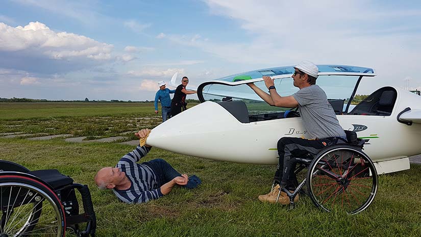 Zdjęcie wykonane na otwartej przestrzeni. Na trawie po prawej stronie widać przednią część szybowca. Przed nim stoi młody chłopak na wózku, który uchyla kabinę. Pod dziobem leży dorosły mężczyzna z nogami amputowanymi poniżej kolan. Leży na prawym boku i lewą ręką wyciera kadłub maszyny.