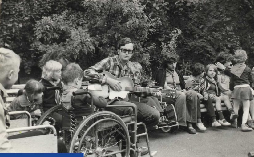 Archiwalne, czarno - białe zdjęcie. Na ławce zwrócony do obiektywu siedzi młody mężczyzna w ciemnych okularach. Gra na gitarze. Po jego lewej stronie widać dzieci na dużych, pokojowych wózkach. Po prawej inne dzieci siedzą na ławkach.