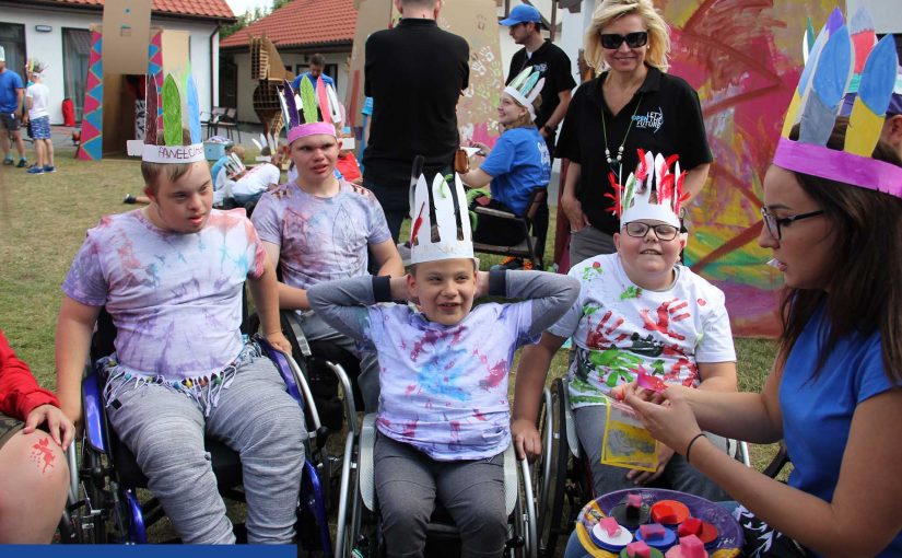 Na zdjęciu czterech chłopców na wózkach w wieku ok. 10-12 lat. Mają na sobie pomalowane na kolorowo T-shirty, a na głowach tekturowe pióropusze. Są bardzo zadowoleni. W tle widać kolorowe tipi również z tektury.