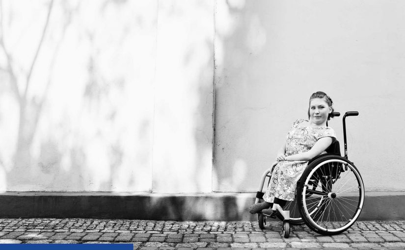 Zdjęcie czarno białe. Po prawej stronie młoda, drobna dziewczyna siedząca na wózku inwalidzkim (widać lewy bok wózka). Dziewczyna zwrócona jest do obiektywu i lekko się uśmiecha. Ręce ma złożone na kolanach. Wózek stoi na ciemnej kostce brukowej, na tle gładkiego muru, na którym widać tylko lekkie cienie drzew.