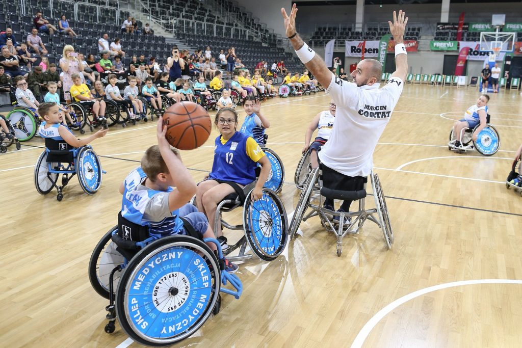 Mecz koszykówki na wózkach. Na boisku odwrócony tyłem z podniesionymi rękoma jest Marcin Gortat (również siedzi na wózku), a wokół niego dzieci. Chłopiec z lewej strony trzyma piłkę do koszykówki nad głową - ujęcie tuż przed rzutem.