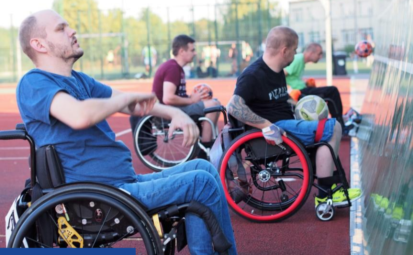 Zdjęcie zrobione na boisku z czerwonym tartanem. Widać czterech mężczyzn na wózkach inwalidzkich, którzy stoją bokiem. Po prawej stronie widoczna ściana, o którą odbijają piłki.